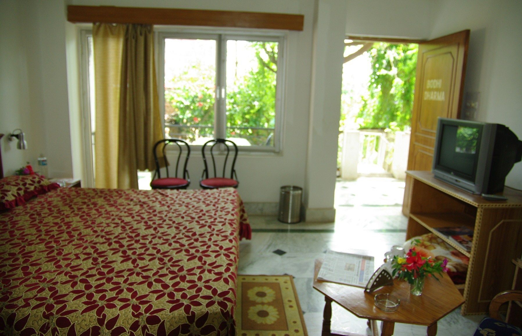 A hotel room with a bed, a television, a small table with a vase, and a window with curtains.