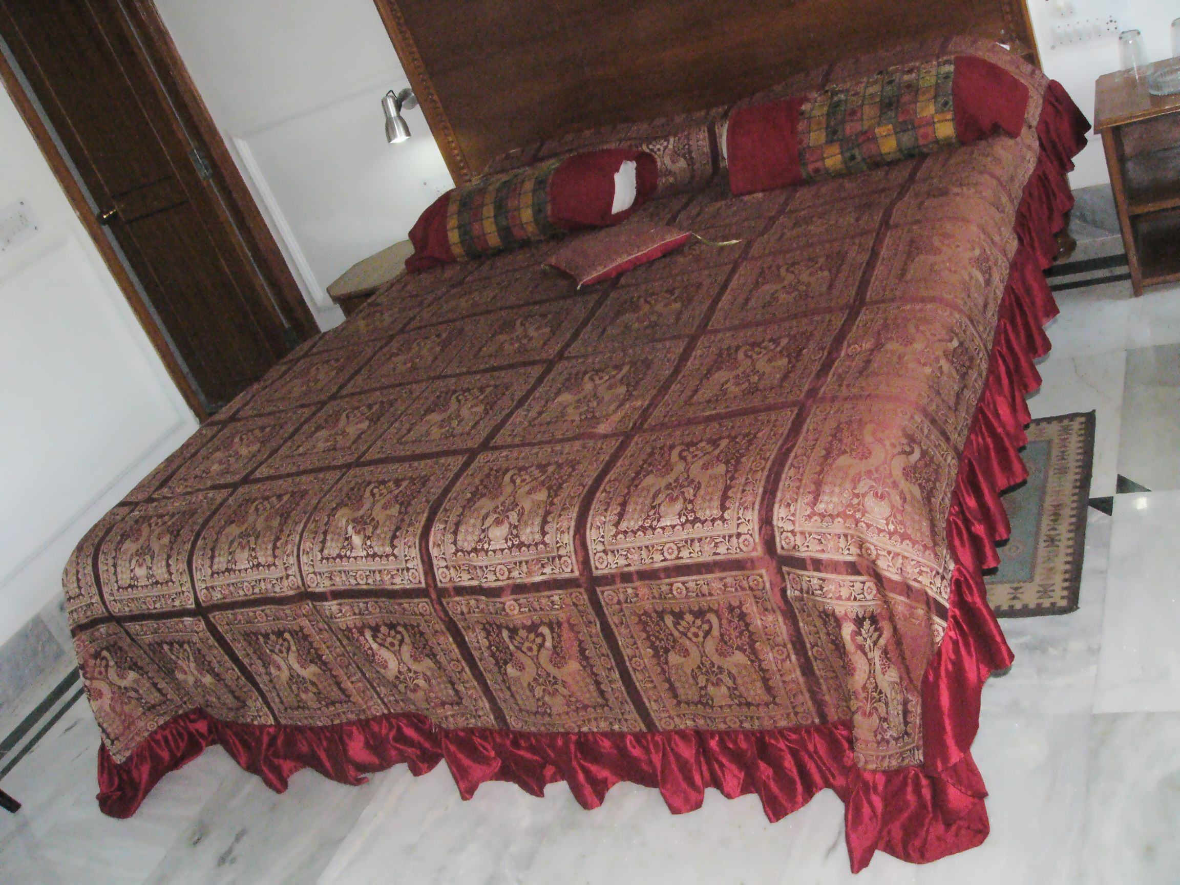A bed with a patterned bedspread and red ruffled edges, a wooden headboard, and a bedside table with a lamp.