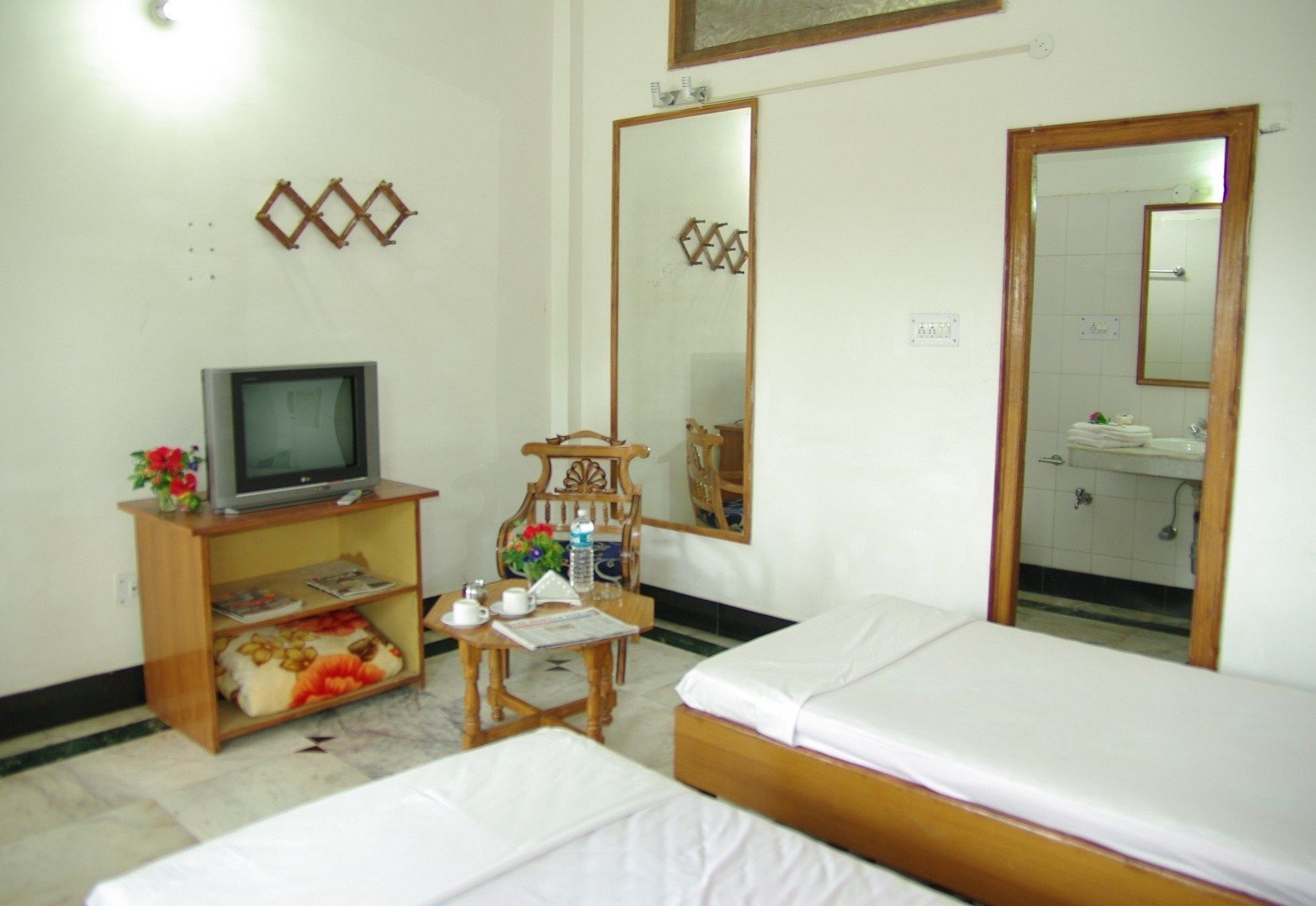 A hotel room with two beds, a television on a wooden stand, a chair, a small table with a newspaper, and a bathroom visible through a mirror.