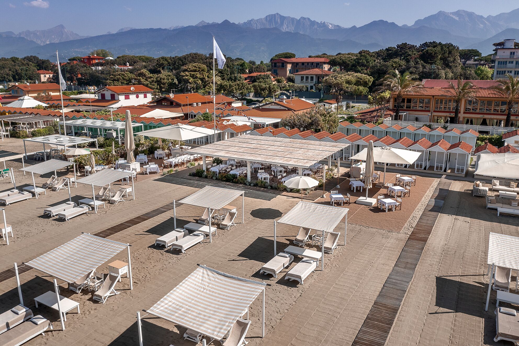 Un hotel sulla spiaggia con numerosi lettini e ombrelloni, una zona pranzo all'aperto coperta e vista sulle montagne sullo sfondo.