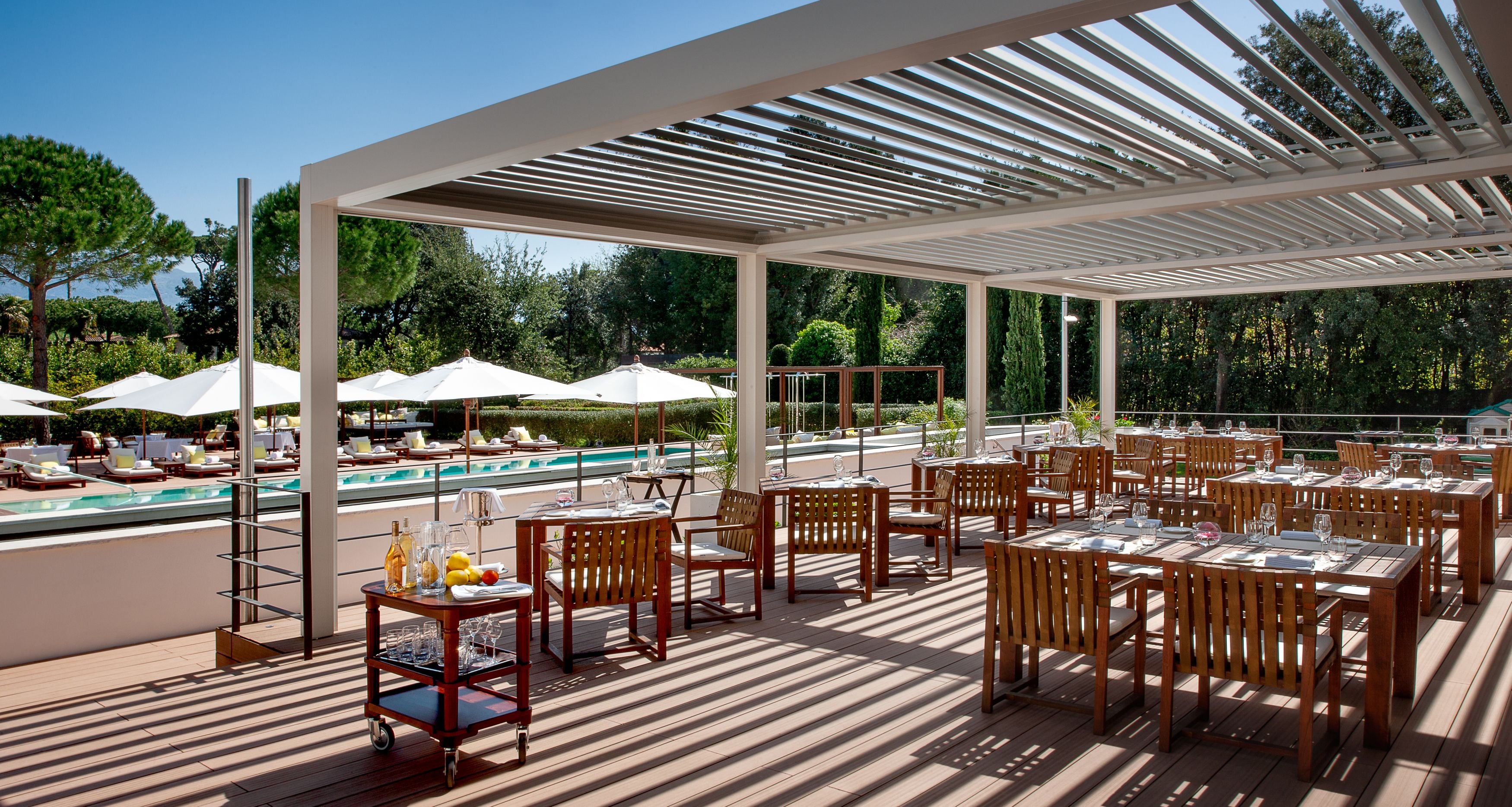 Zona pranzo all'aperto con tavoli, sedie, piscina e ombrelloni.