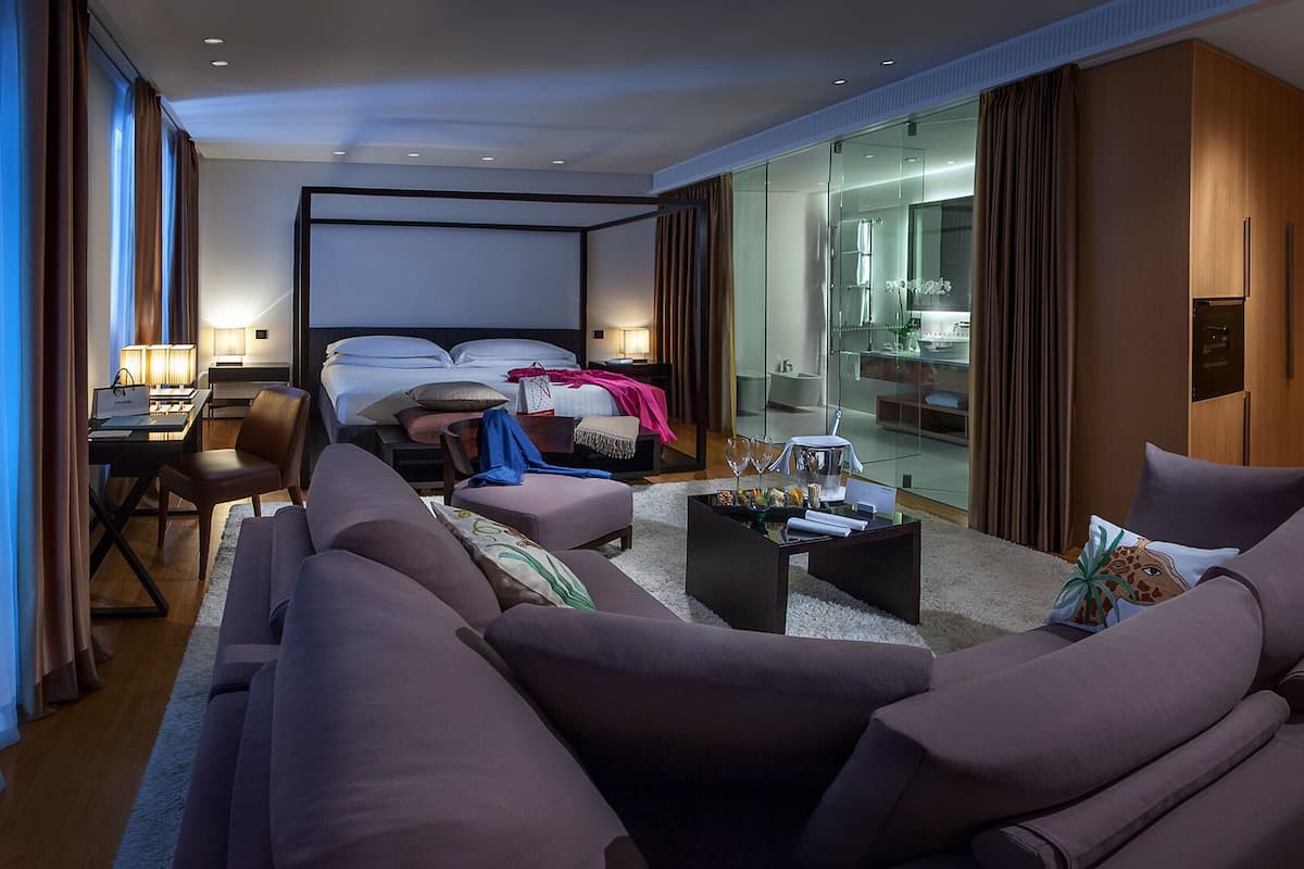 A modern hotel room with a large bed, a seating area with a sofa and armchairs, a small table with a lamp, and a glass-enclosed bathroom visible through the glass door.