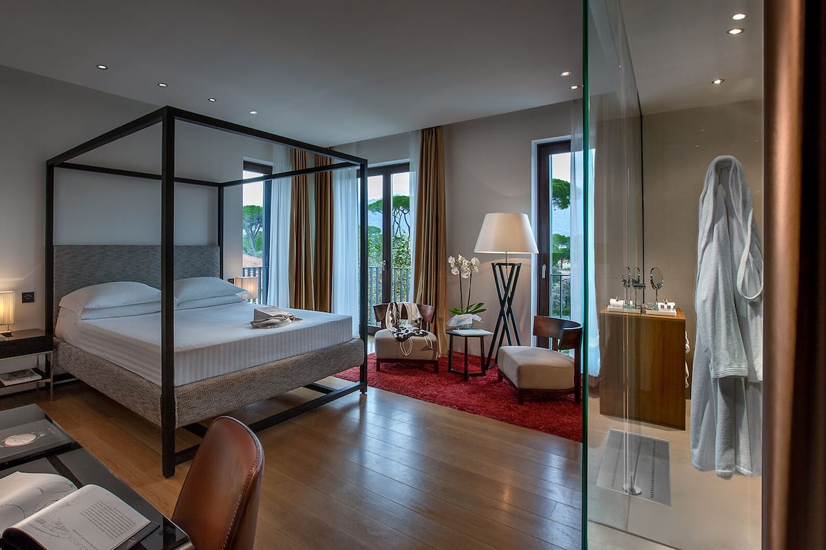 A modern hotel room with a four-poster bed, a desk with a lamp, a chair, and a red carpet.