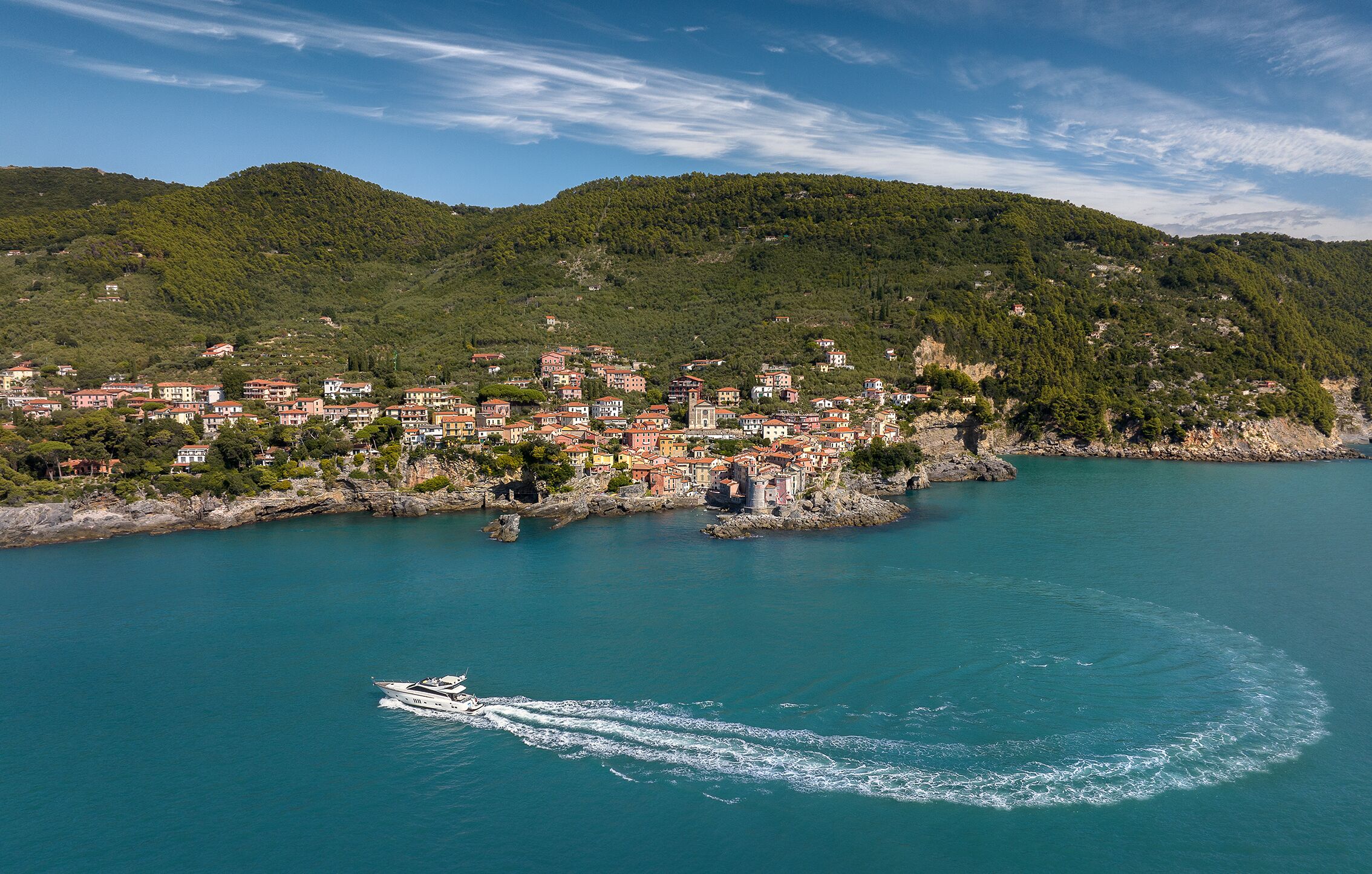 A coastal town with buildings along the shoreline, a boat creating a wake, and a mountainous backdrop.