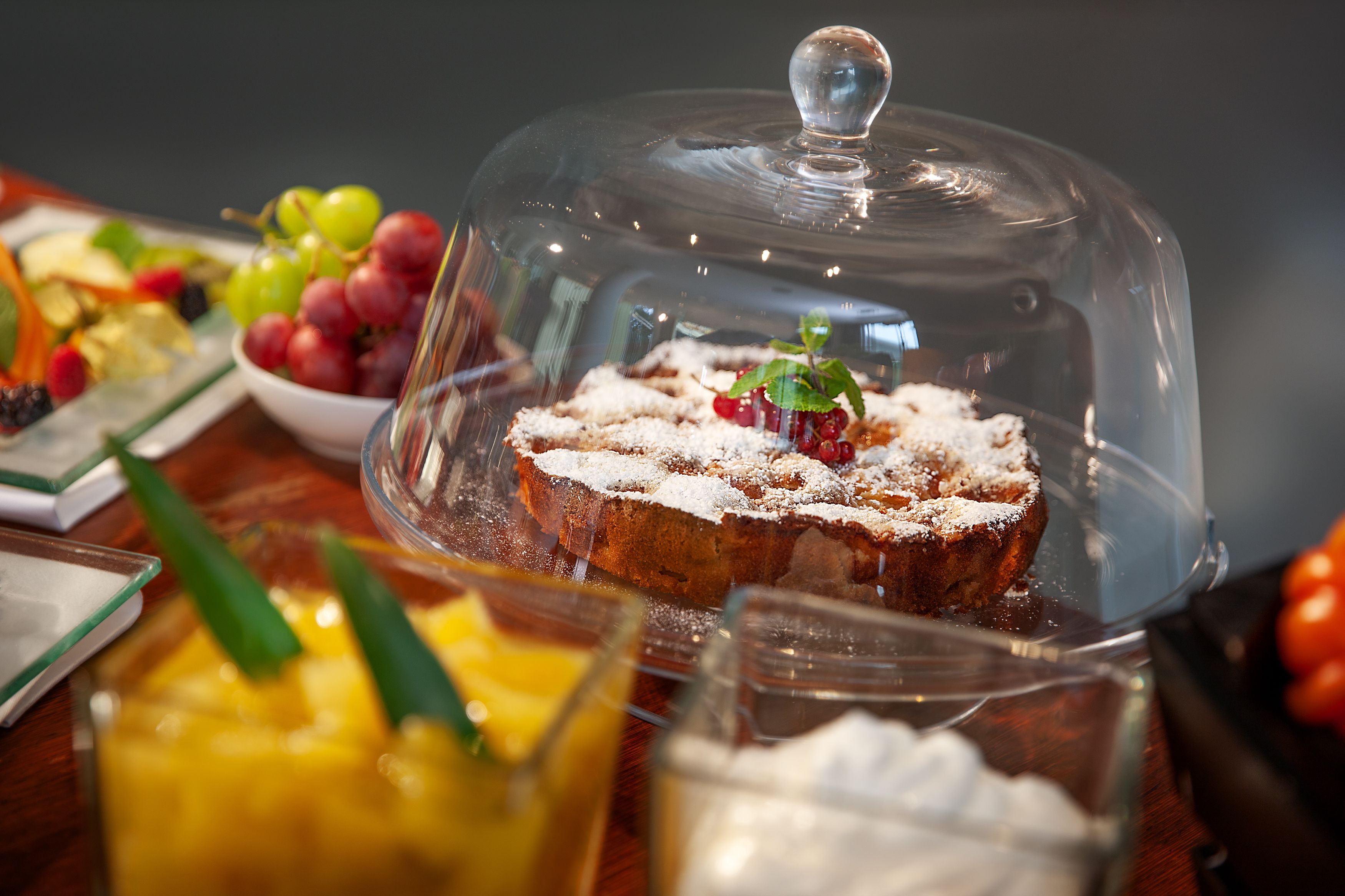 A dessert covered with a glass dome, accompanied by a bowl of mixed fruits and a glass of a yellow beverage.