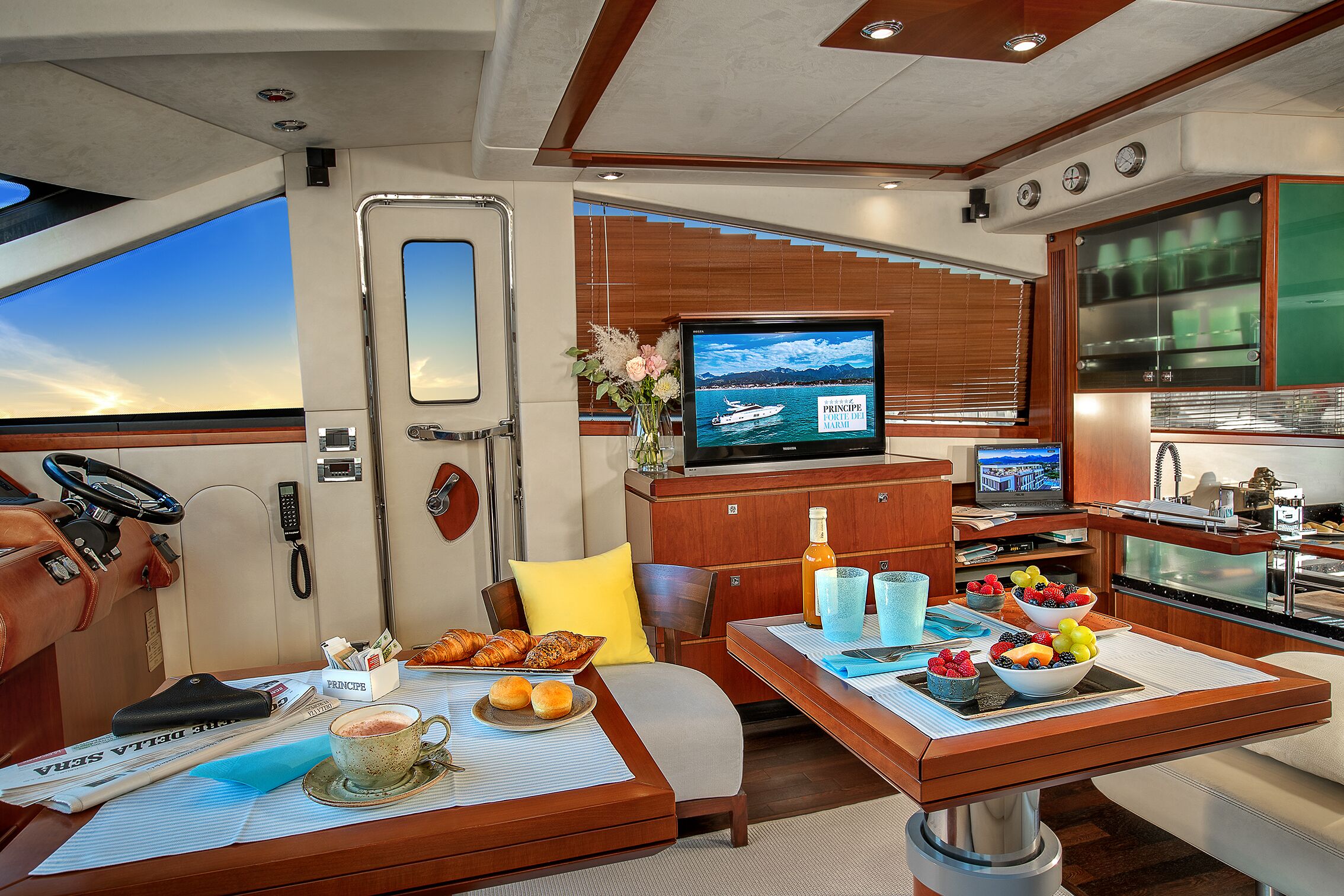 A yacht interior with a dining area, a TV displaying a boat, and a kitchen area.