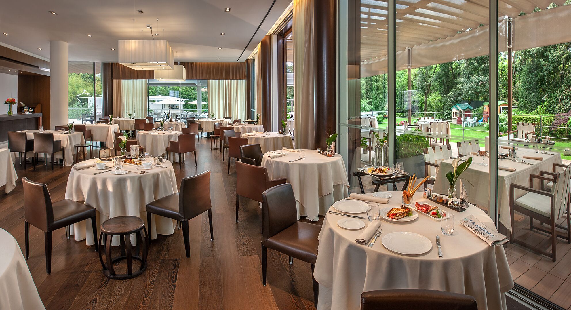 Un restaurante espacioso con mesas y sillas redondas, ventanas amplias que ofrecen vistas al exterior y una decoración de diseño moderno.