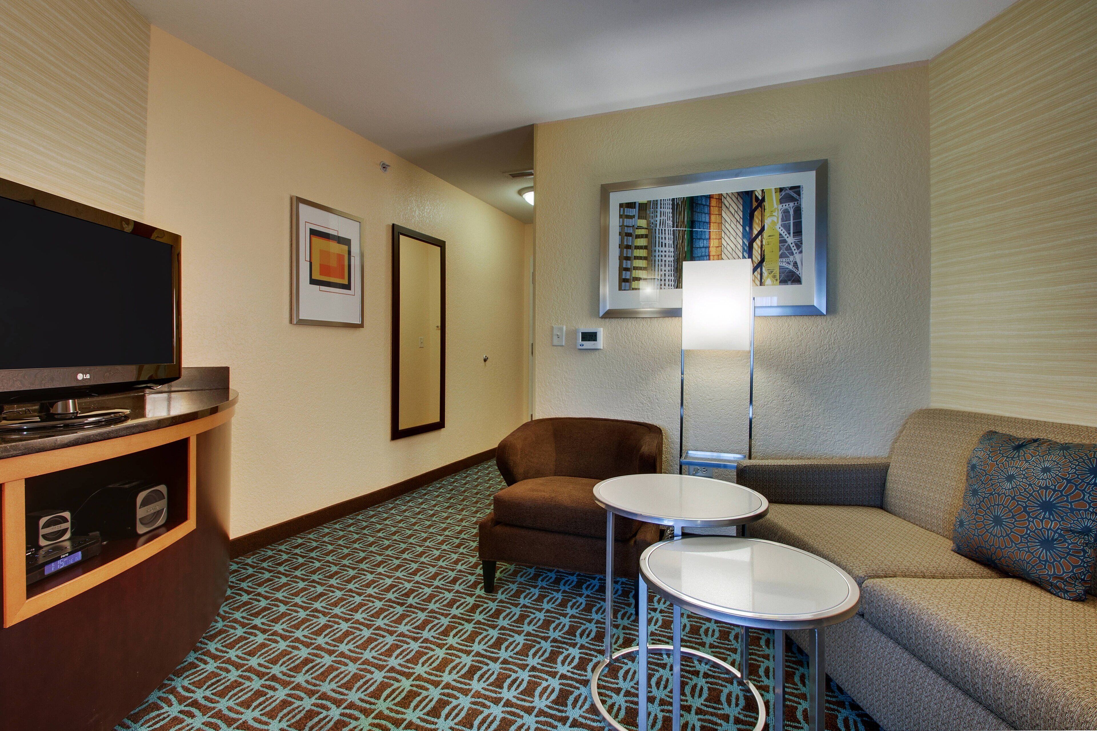 Habitación de hotel con una televisión de pantalla plana, un sillón marrón, un sofá, una mesita redonda pequeña y una alfombra estampada.