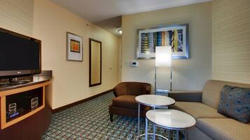 Habitación de hotel con una televisión de pantalla plana, un sillón marrón, un sofá, una mesita redonda pequeña y una alfombra estampada.
