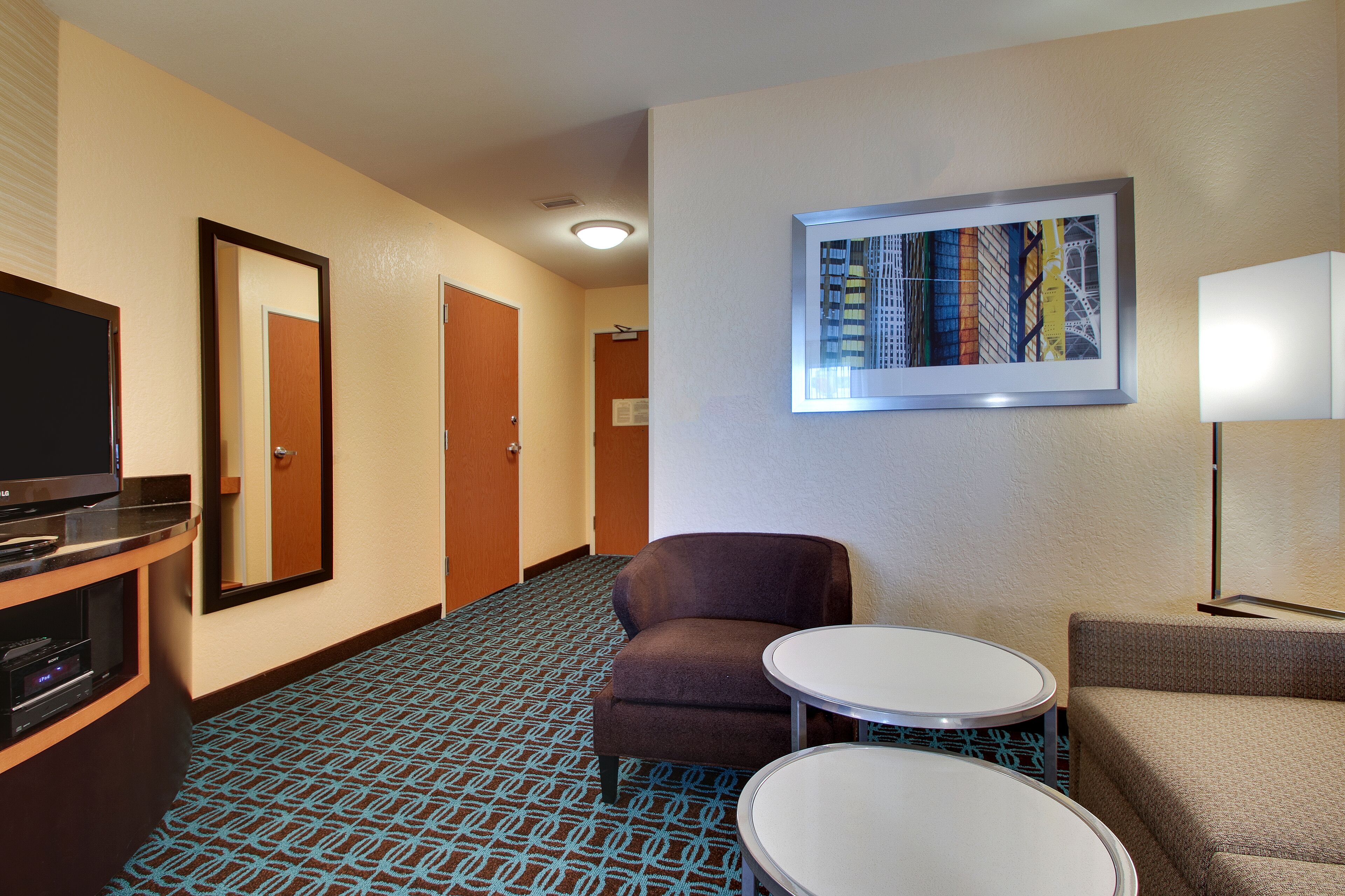 Une chambre d’hôtel avec une moquette à motifs, une télévision, un canapé, une petite table et un tableau encadré au mur.