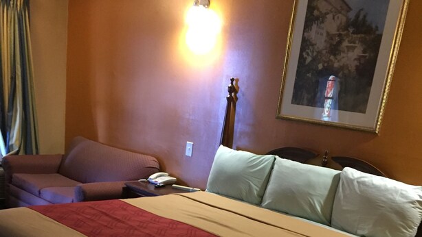 A hotel room with a bed, a sofa, a nightstand, a lamp, and a painting on the wall. - Budget Inn And Suites (Abbeville)