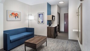A modern hotel room with a blue sofa, a wooden coffee table, and a kitchenette area. - Holiday Inn Express Hotel & Suites Pratt by IHG (Pratt)