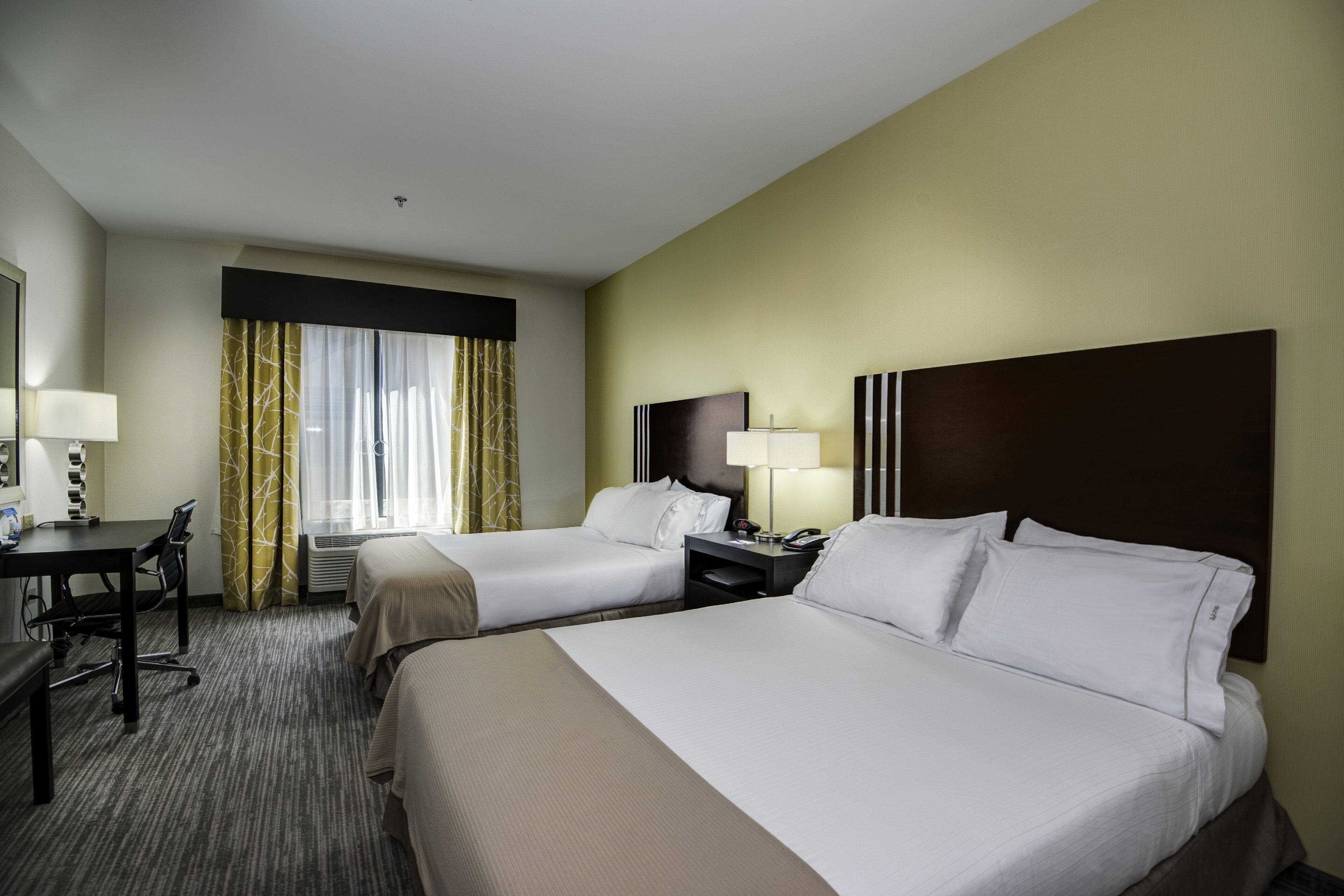A hotel room with two beds, a desk, a chair, a lamp, and a window with curtains.