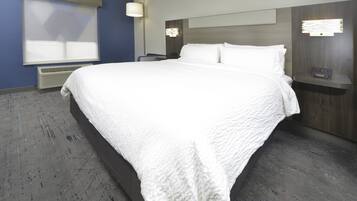 A neatly made bed with white linens in a hotel room with a blue accent wall, a wall-mounted heater, and a bedside table with a lamp.