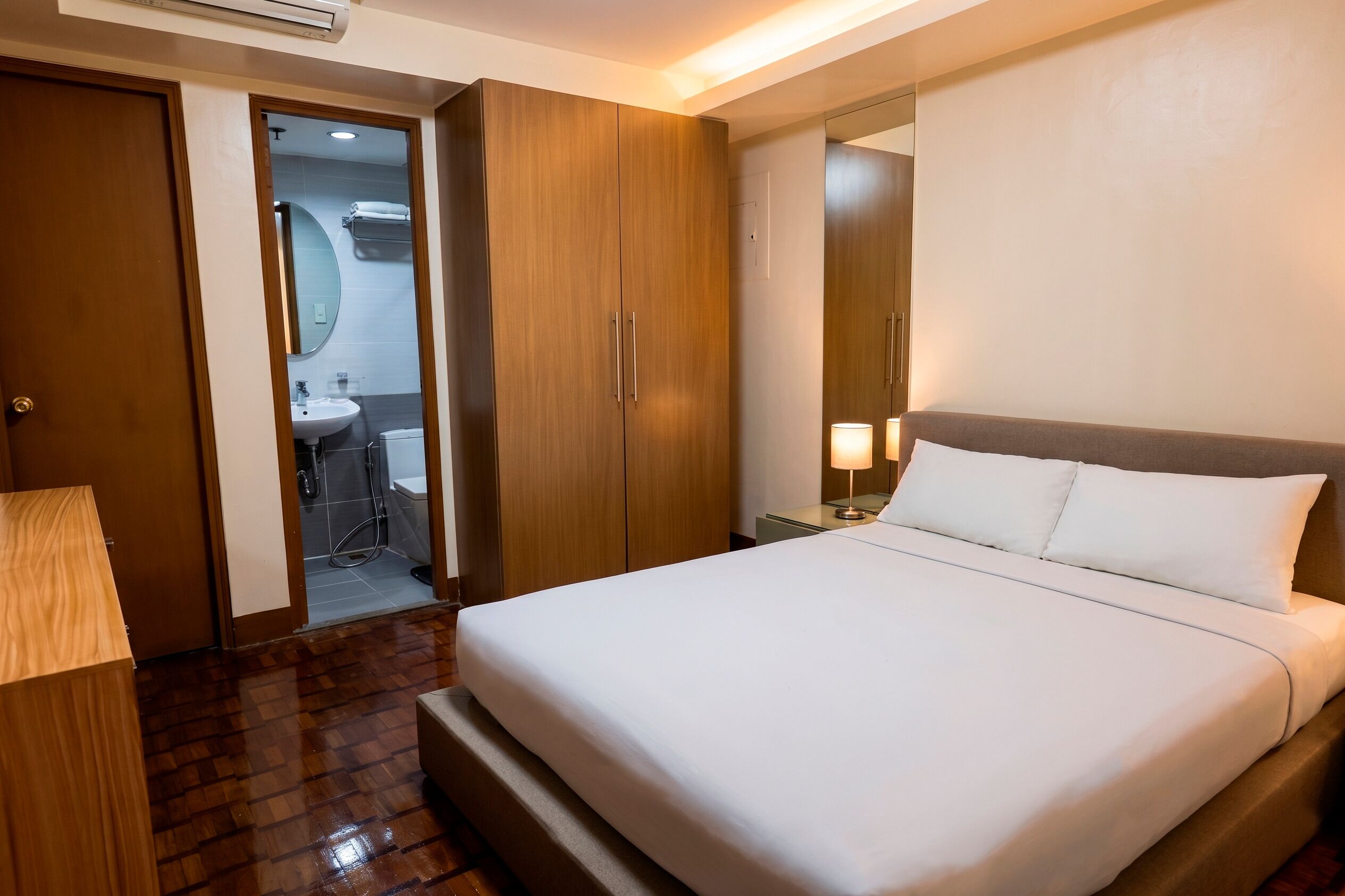 A hotel room with a bed, a nightstand, a lamp, a wooden wardrobe, and a bathroom visible through an open door.