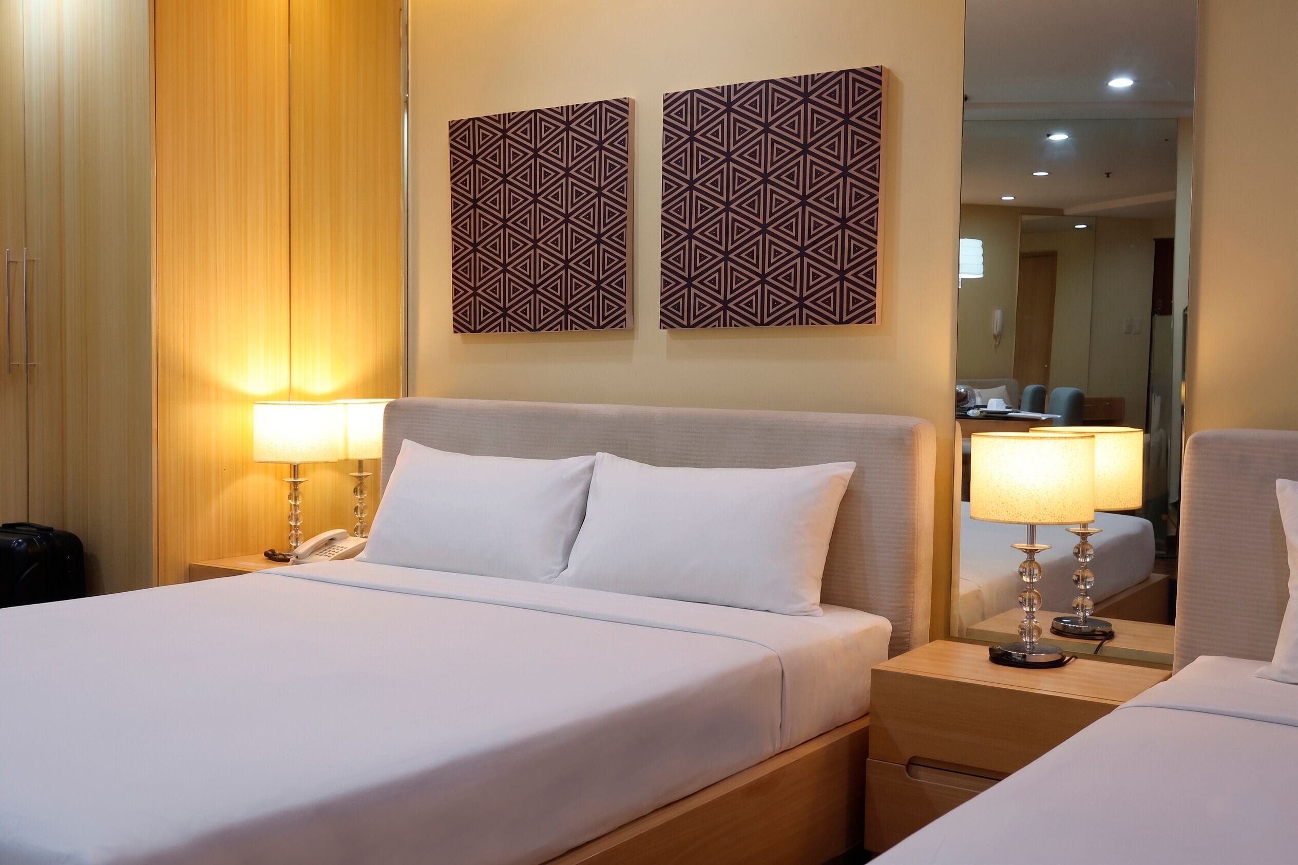 A hotel room with a bed, bedside lamps, a telephone, a mirror, and two wall-mounted decorative panels.