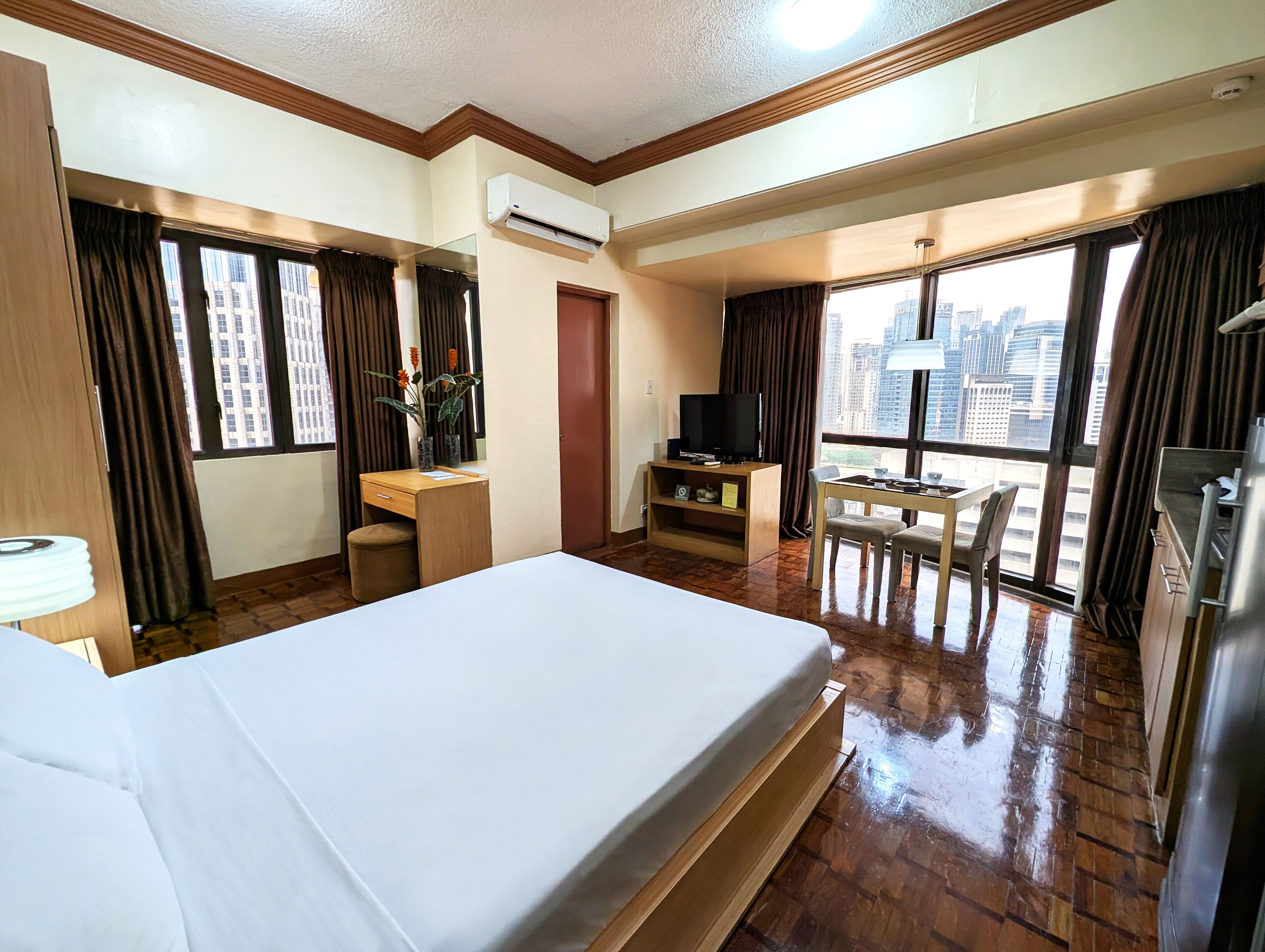 Ein Hotelzimmer mit Bett, Schreibtisch, Fernseher, einem kleinen Tisch mit Stühlen und Blick auf die Stadt durch große Fenster.