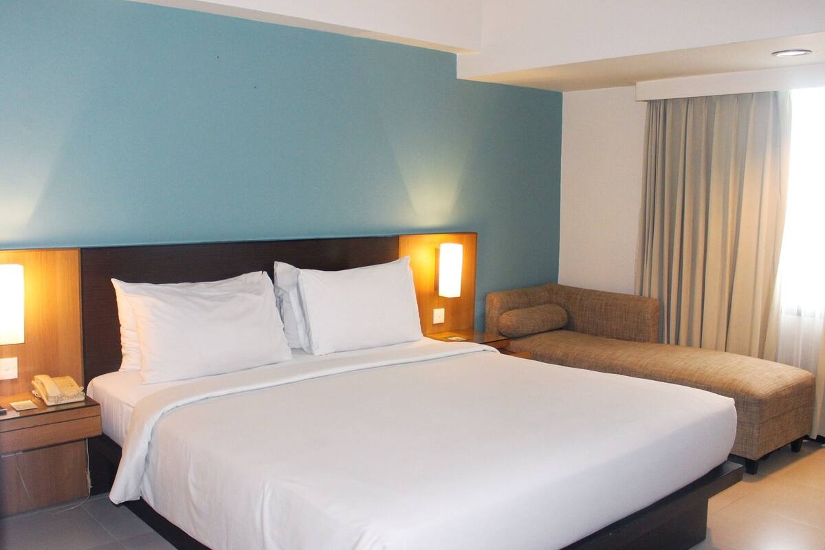 A hotel room with a large bed, a bedside table with a lamp, a telephone, and a sofa.