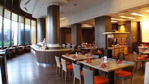 Un restaurante moderno con una barra central, varias mesas de comedor con cojines naranja y grandes ventanales que dejan entrar luz natural.