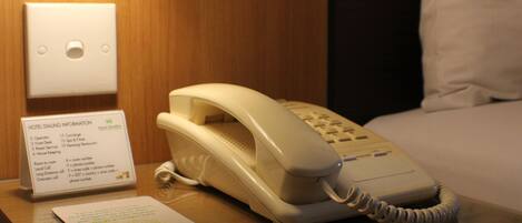 A hotel room with a bedside telephone, a sign with hotel contact information, and a glass table.