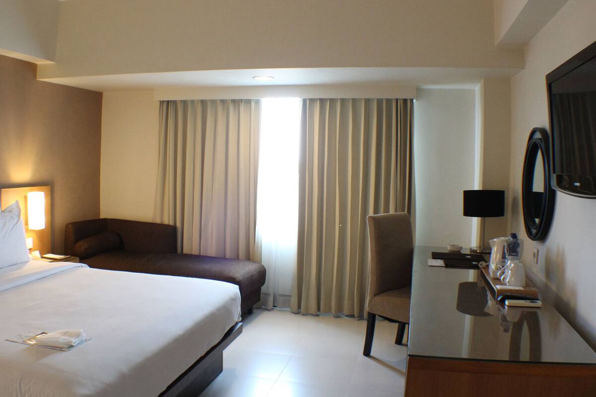 A hotel room with a large bed, a desk with a chair, a mirror, and a window with curtains.