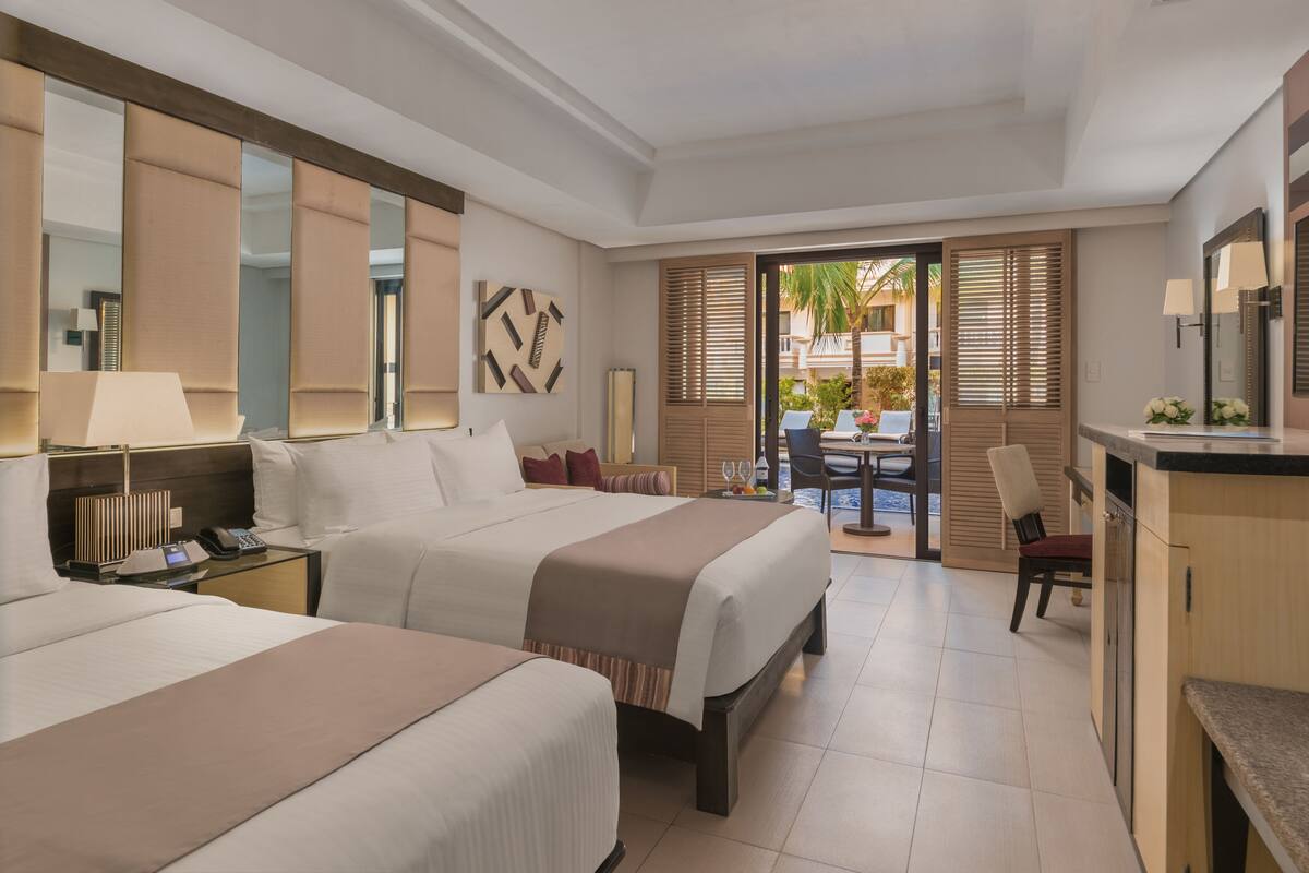 Habitación de hotel con dos camas, un escritorio, una silla, una mesa de comedor y un balcón con vistas a palmeras.