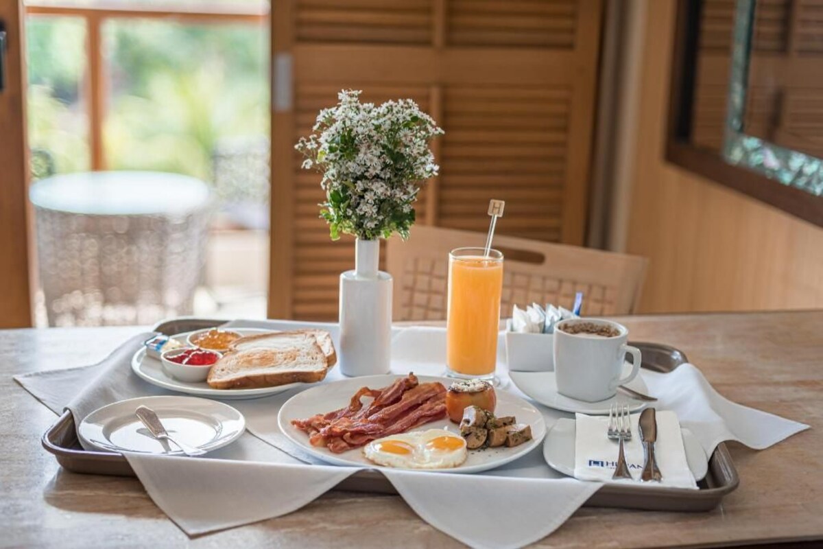 Bandeja de desayuno con tostadas, huevos, tocino y un vaso de jugo de naranja sobre una mesa con un florero.