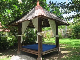 A wooden gazebo with a thatched roof, a blue mattress, and white curtains.