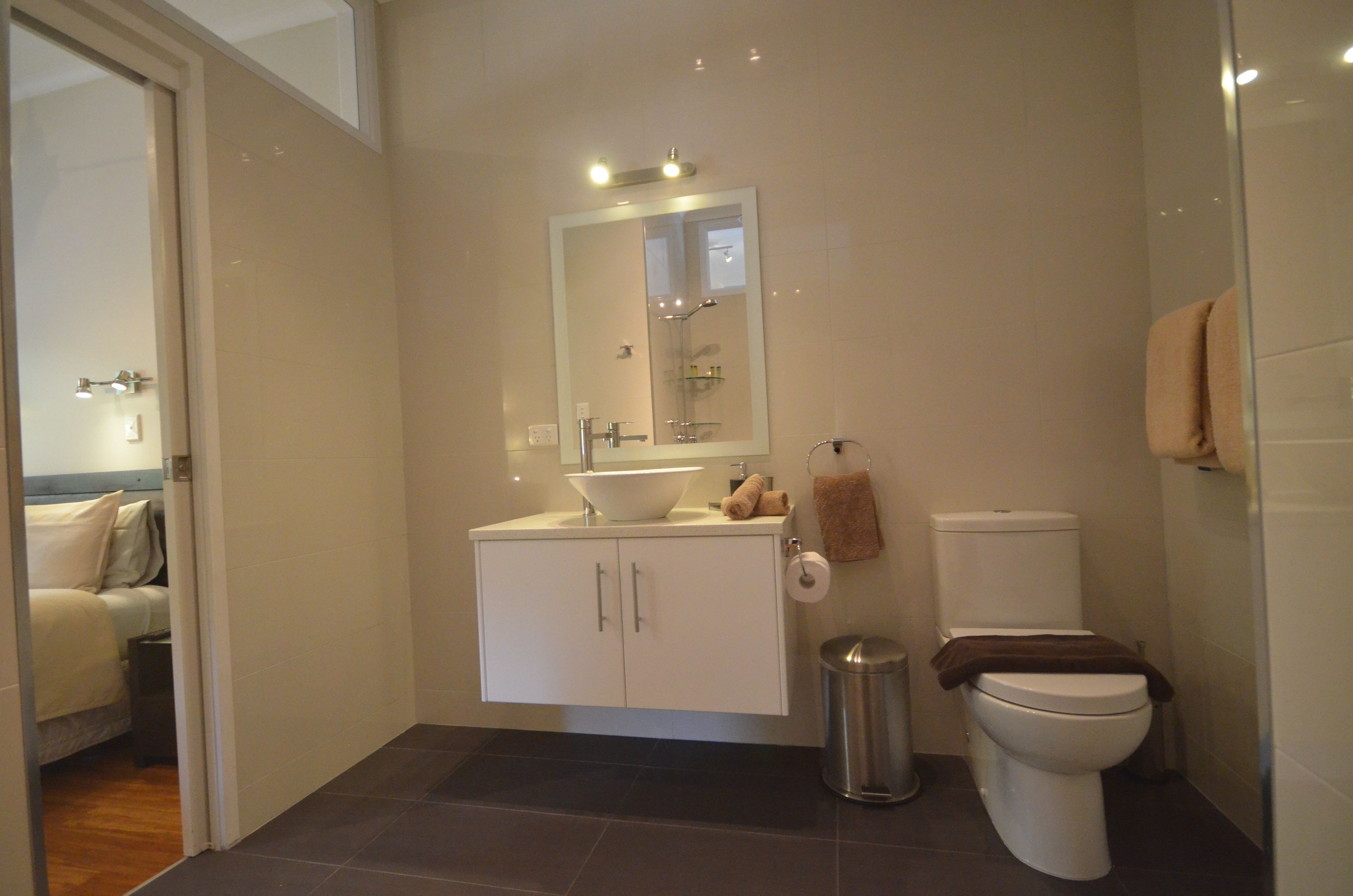 Un baño moderno con lavamanos, espejo y retrete blancos. Se ve un cubo de basura y un perchero para toallas.