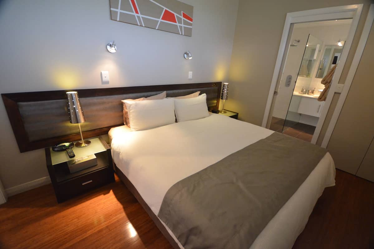 Una cama bien tendida con cabecera de madera, una mesita de noche con lámpara, y un espejo en la habitación contigua.