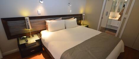 A neatly made bed with a wooden headboard, a bedside table with a lamp, and a mirror in the adjacent room.