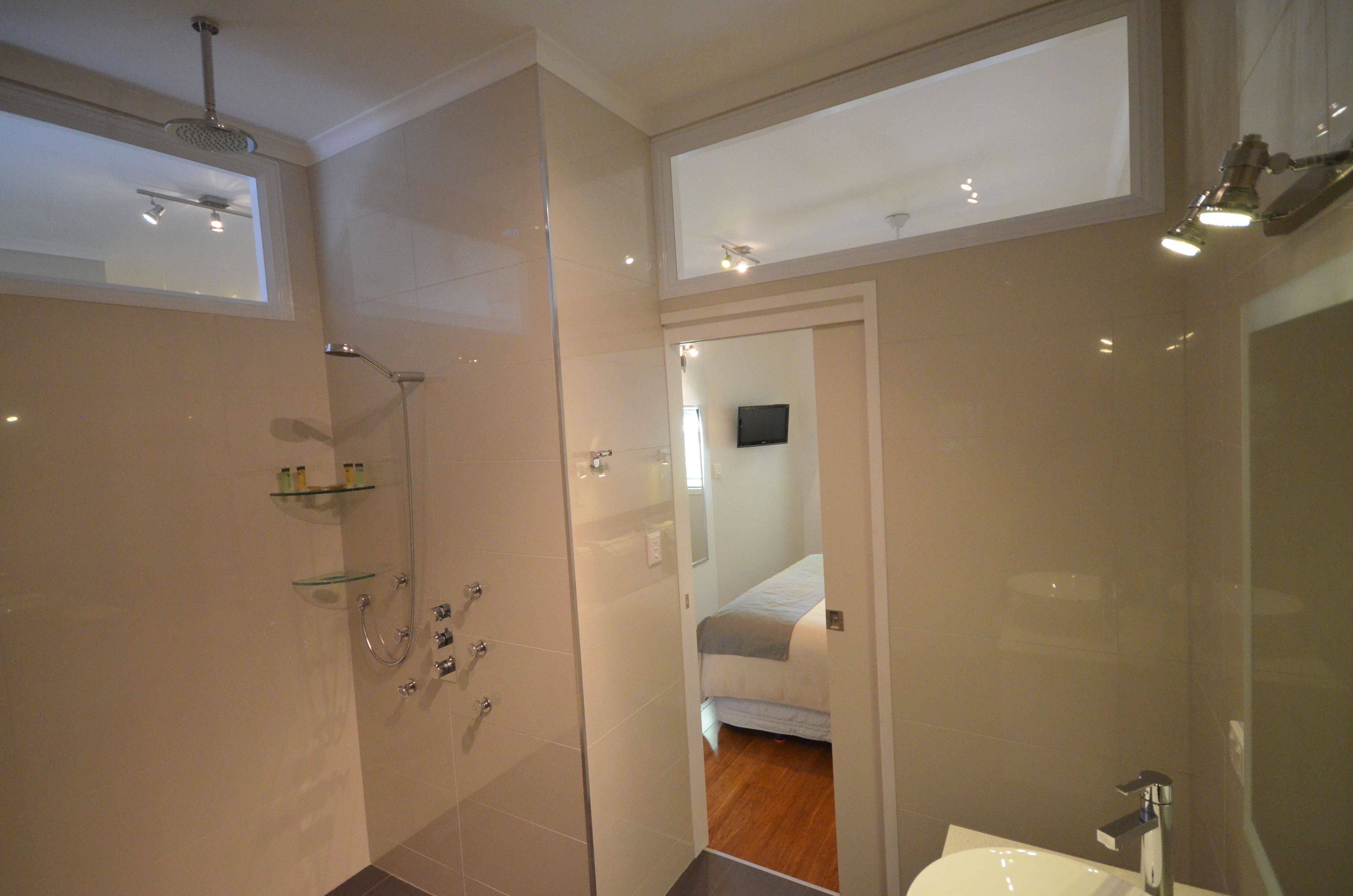 A modern bathroom with a glass shower enclosure, a visible bed in the adjacent room, and a mounted TV.