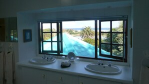 A bathroom with two sinks, a mirror, and a window with a view of a beach and palm trees. - Mont Fleur B&B (Wilderness)