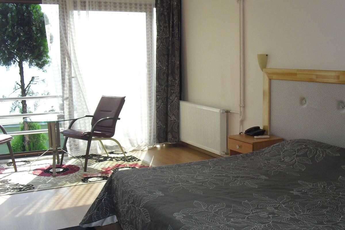 A bedroom with a bed, a chair, a bedside table, a radiator, and a window with curtains.