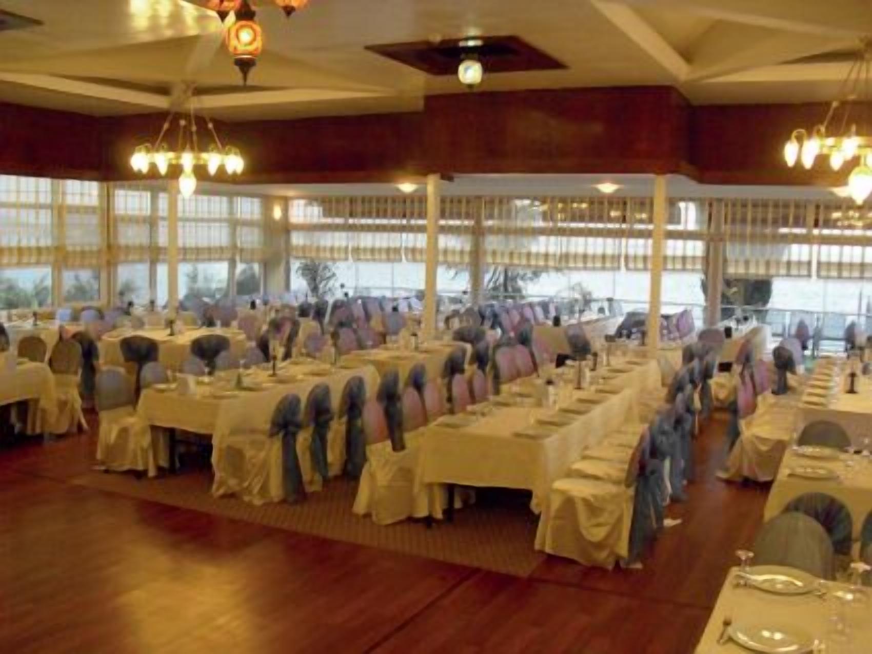 Un salón de banquetes con filas de mesas dispuestas para un evento formal, ventanas amplias y candelabros.