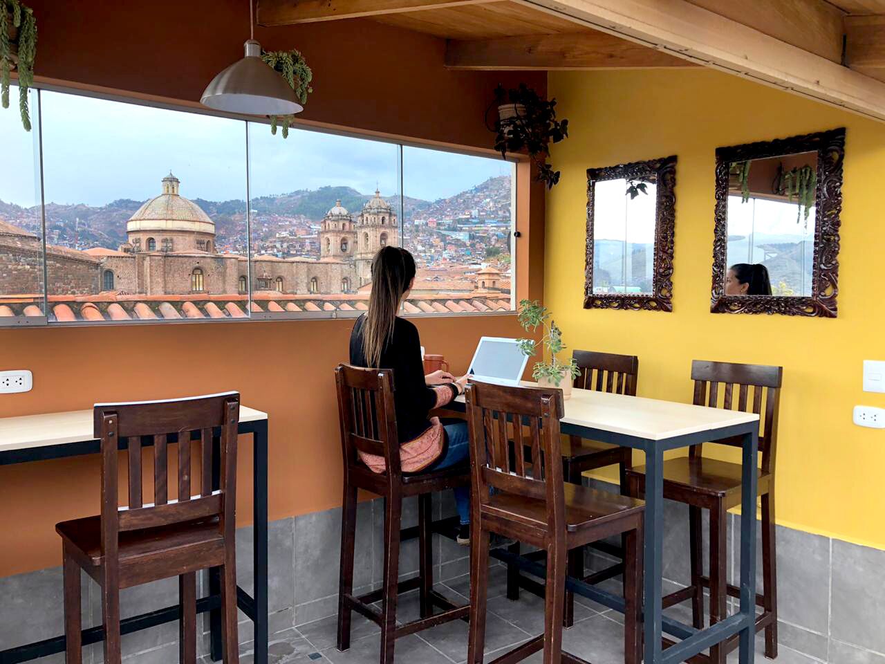 Une femme est assise à une table entourée de chaises et utilise un ordinateur portable. La table est placée devant une fenêtre donnant sur un paysage urbain avec des bâtiments historiques.