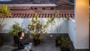 A person meditating on a green mat on a rooftop patio with potted plants, overlooking a cityscape with terracotta rooftops and a mountain in the background. - Maytaq Wasin Hotel (Cusco)