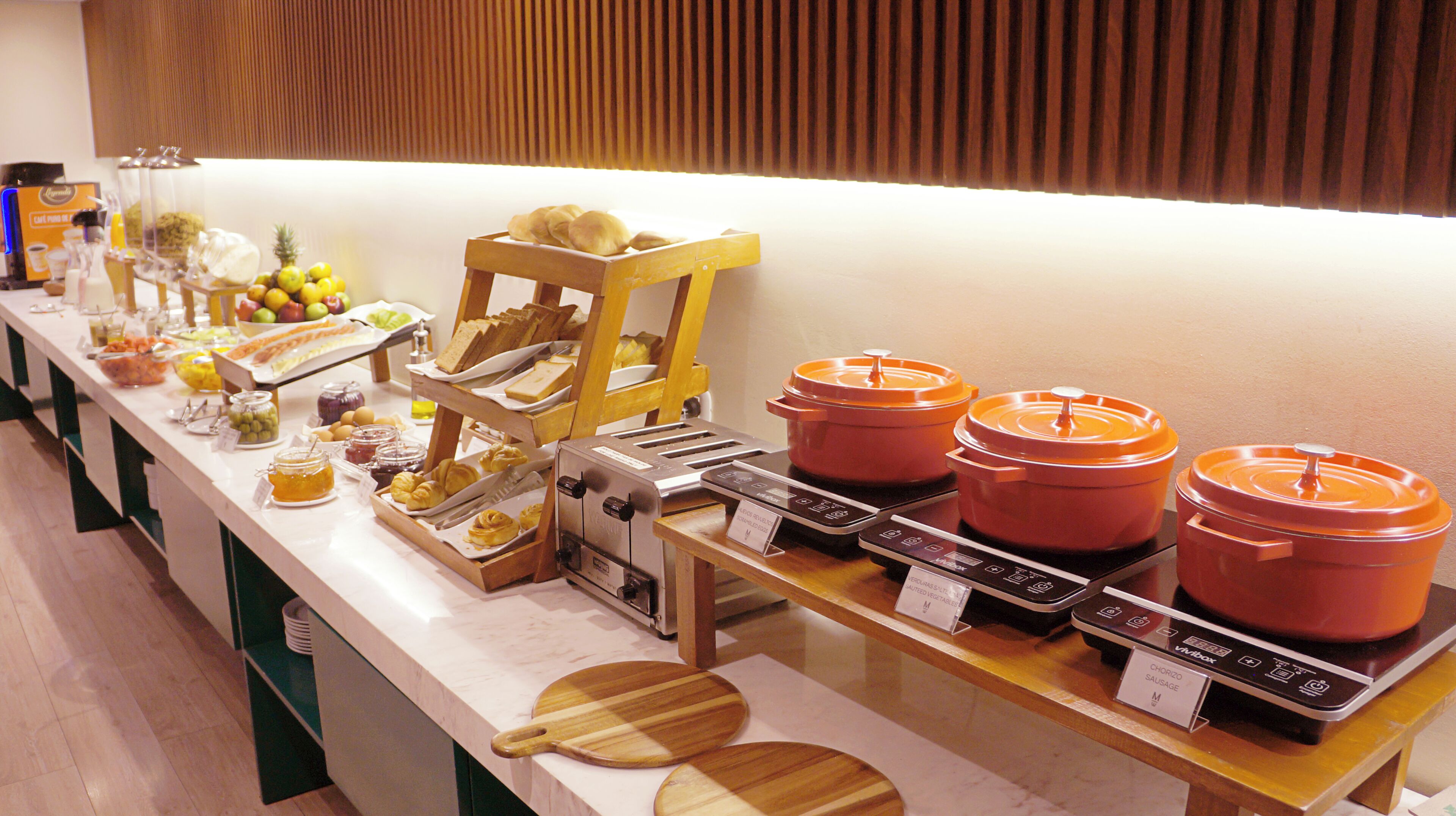 Un bufé con diferentes opciones de comida, incluyendo frutas, pastelería y platos calientes, dispuesto en una mesa larga.