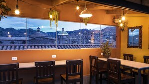 A dining area with a large window overlooking a cityscape, featuring a table and chairs, hanging lights, and a potted plant. - Maytaq Wasin Hotel (Cusco)