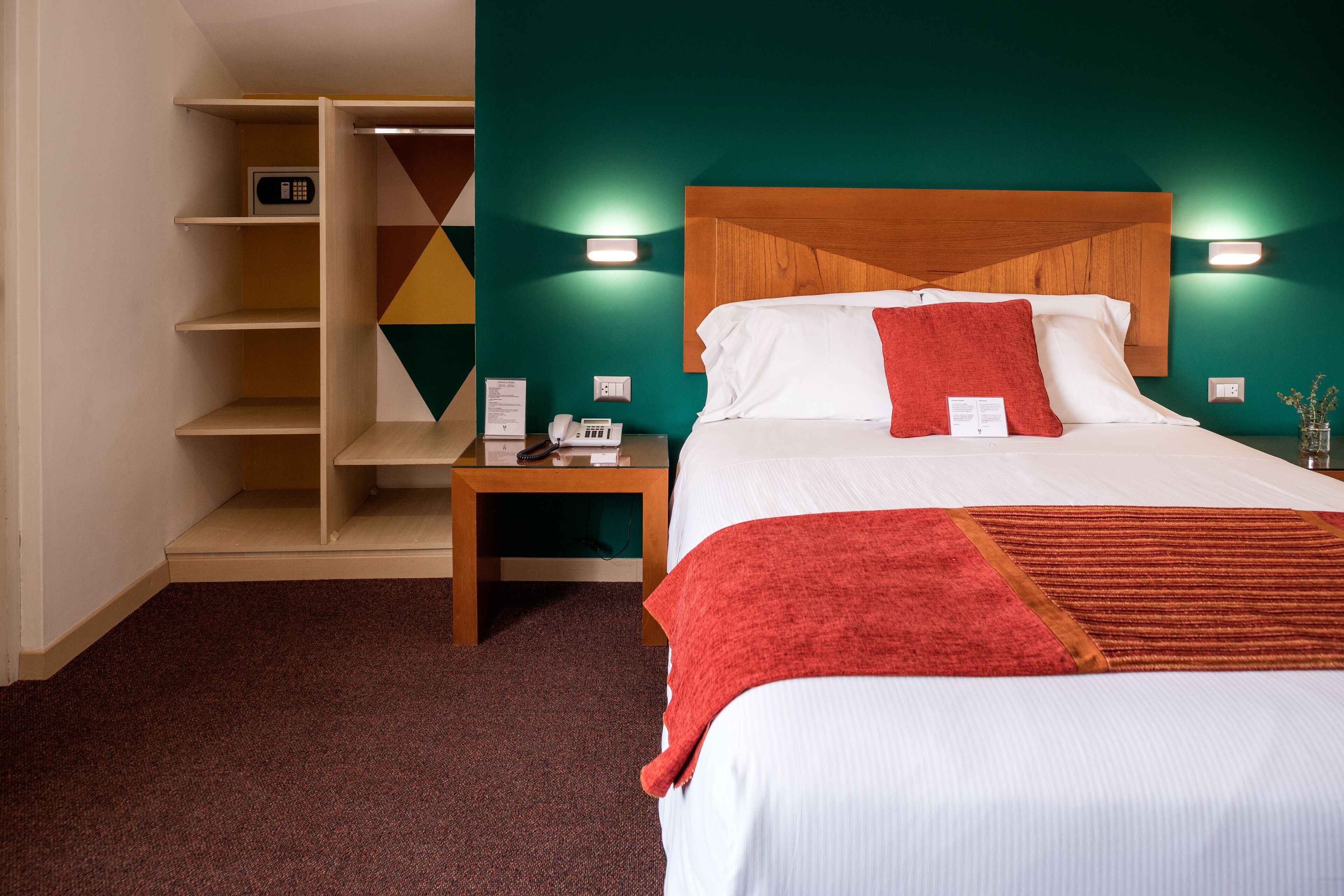 A hotel room with a bed, a nightstand, a desk with a telephone, and a storage unit with shelves.
