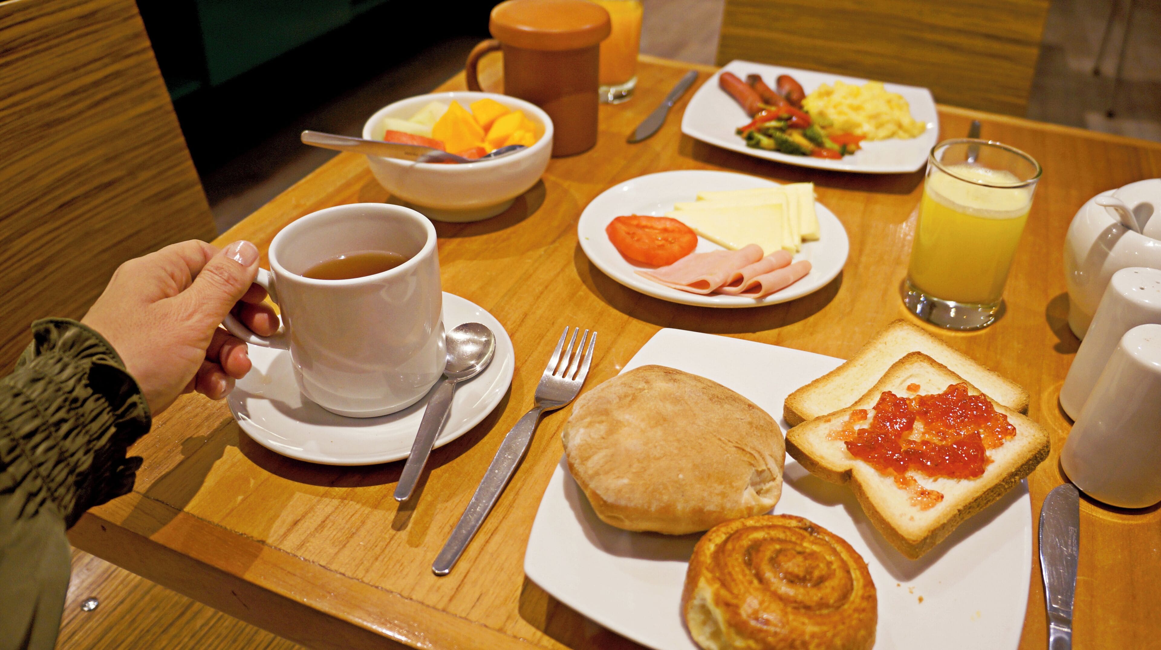 Una mesa de desayuno con una taza de café, un plato de huevos, salchichas, tostadas con mermelada, un muffin y un bol de fruta.