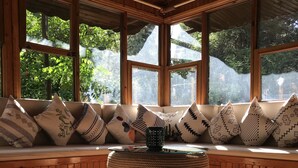 Un coin salon accueillant avec un banc en bois et des coussins, un pouf en osier et de grandes fenêtres donnant sur un paysage verdoyant.