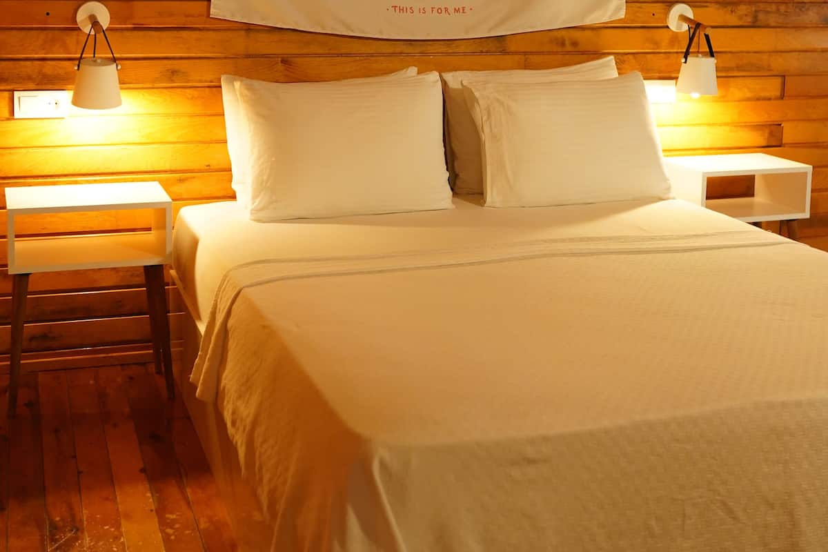 Een bed met witte beddengoed, twee nachtkastjes met lampen, en een houten wand met wandtapijt.