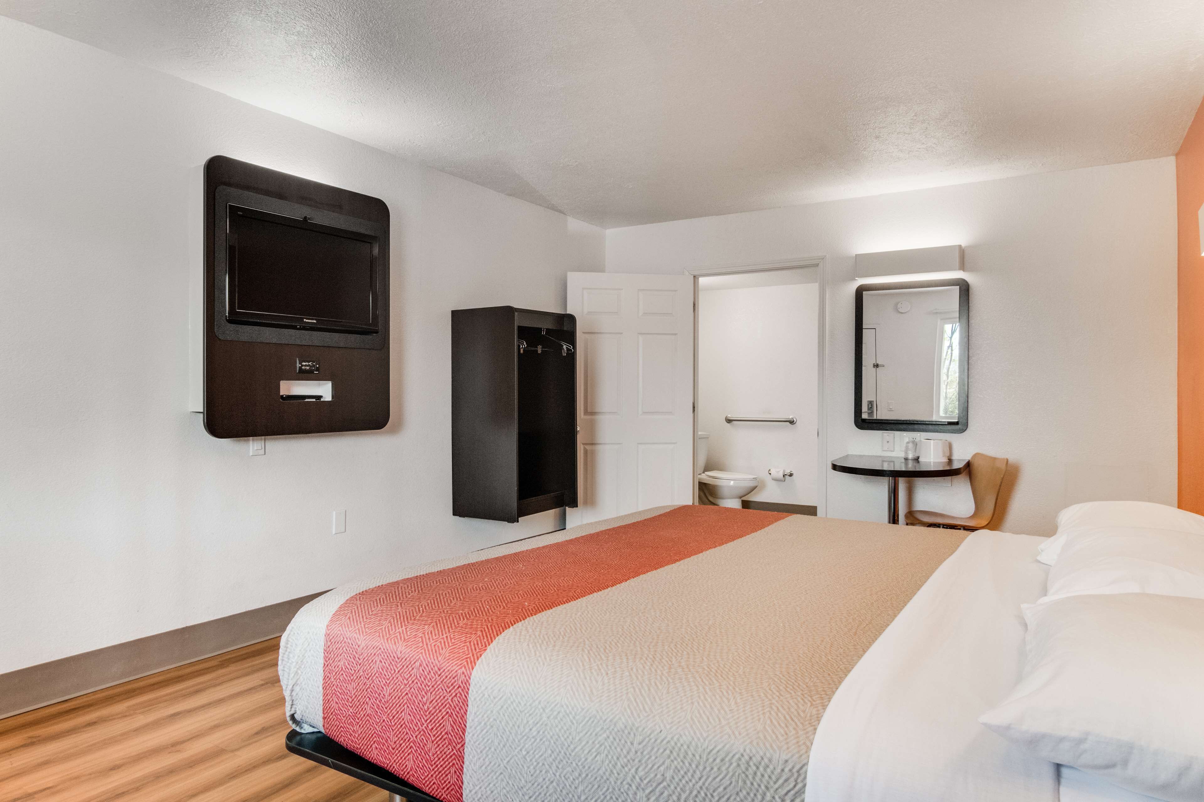 A hotel room with a bed, a TV mounted on the wall, a wardrobe, a small table with a mirror, and a sink.