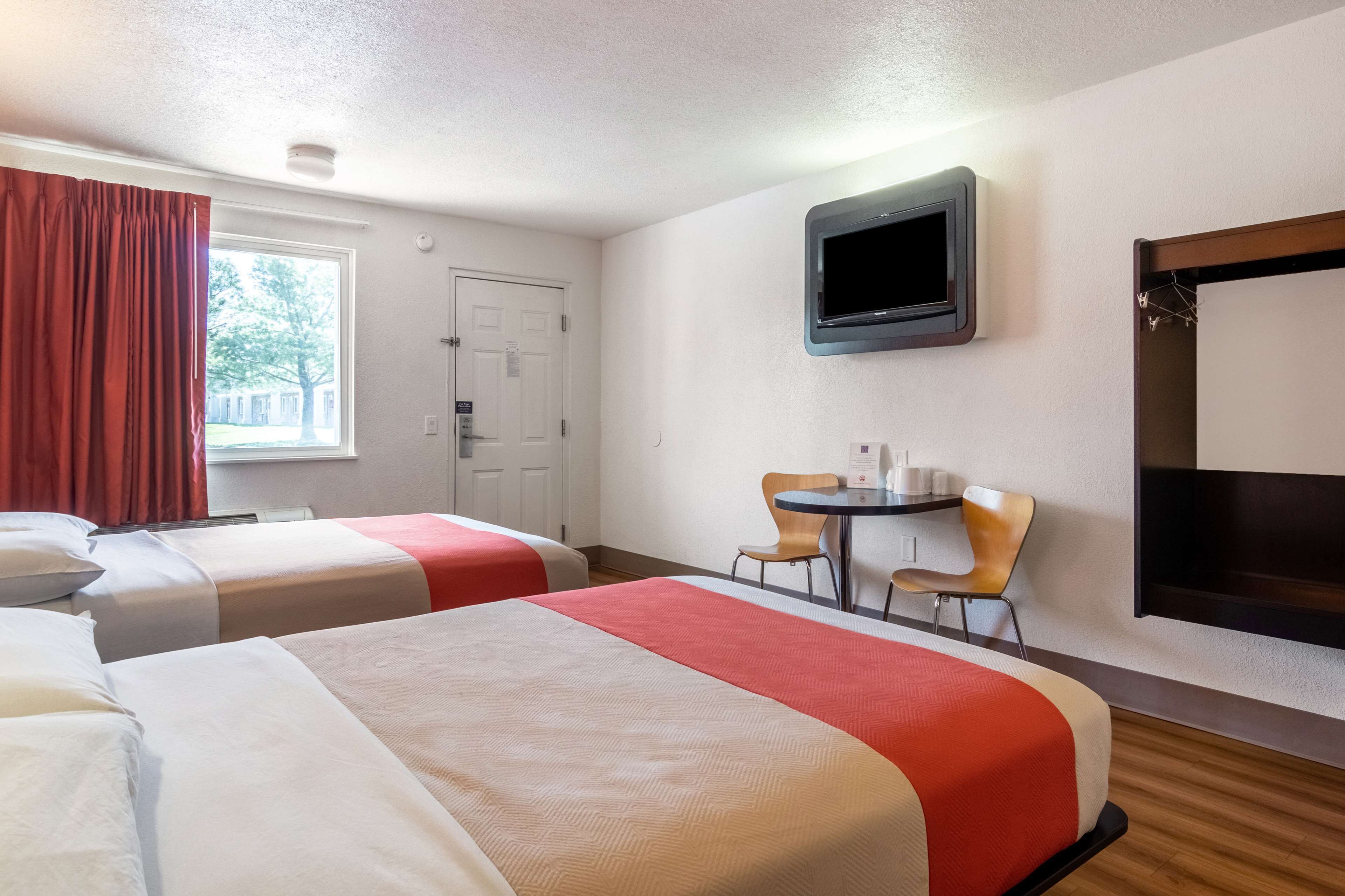 A hotel room with two beds, a TV, a small table with chairs, and a window with curtains.
