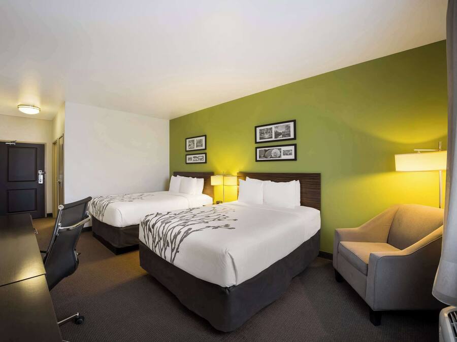 A hotel room with two beds, a desk, a chair, and a lamp.