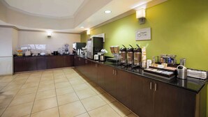 A hotel kitchen with a long counter, equipped with coffee machines, a refrigerator, and various food items.