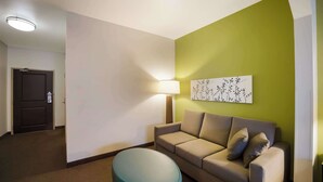 A hotel room with a beige sofa, a teal ottoman, a wall-mounted lamp, and a door in the background.