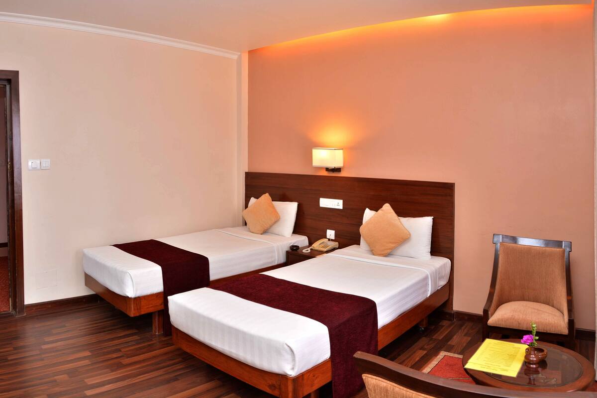 A hotel room with two single beds, a wooden headboard, a nightstand with a phone, a chair, and a small table with a flower vase and a menu.