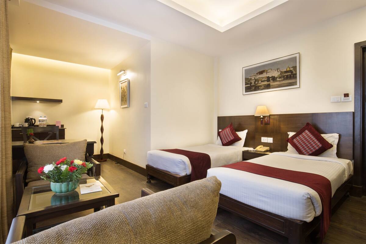 A hotel room with two beds, a desk with a lamp, a chair, a coffee maker, and a vase of flowers.